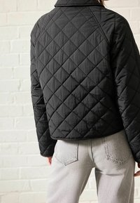Černá prošívaná bomber bunda s krátkým střihem, se hladkým vnějším povrchem a diamantovým vzorem. Šedé džíny viditelné dole.