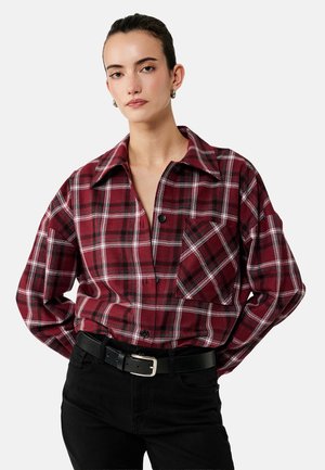 Vrouw draagt oversized rood-zwart geruit shirt met voorzak, zwarte riem en zwarte broek, staand met handen achter de rug.