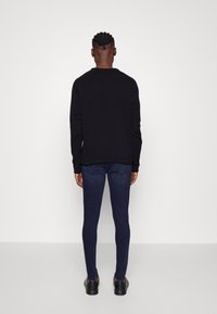 Sweatshirt preto com detalhes canelados, combinado com jeans slim fit azul escuro e sapatos pretos. A vista é de trás, destacando a forma do conjunto.