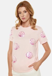 Lyserød bomulds-t-shirt med pailletbesatte muslingedesigns i forskellige størrelser på forsiden. Korte ærmer og rund halsudskæring. Glat tekstur.