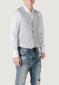 Gilet a righe grigie e bianche con tasche frontali, chiusura a bottoni, indossato sopra una camicia bianca e jeans blu strappati. Texture liscia, vestibilità su misura.