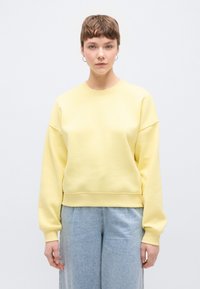 Ανοιχτό κίτρινο φούτερ με άνετη εφαρμογή, στρογγυλή λαιμόκοψη και ραφές ώμων σε χαμηλό σημείο, συνδυασμένο με ανοιχτό μπλε τζιν παντελόνι φαρδιάς γραμμής.