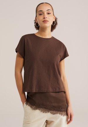 Naine, kellel on rõngas-kujulised kõrvarõngad, kannab pruuni lühikeste varrukatega tuba, mille all on pitsilise äärega pruun kampsun ning helebeeže värvi pükse.