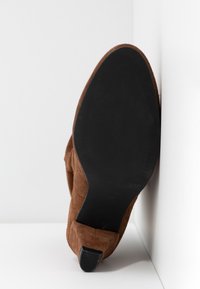 Chaussure en suède marron en angle contre un coin, montrant une semelle noire lisse avec un petit embout de talon en caoutchouc noir.