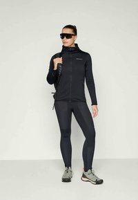 Černá sportovní bunda se zipem, v kombinaci s černými legínami. Modelka nosí sluneční brýle a šedé turistické boty, drží černý batoh.