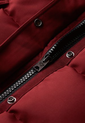 Bordeaux pufferjack die beschikt over een zwarte rits, zilveren drukknopen en een gewatteerde textuur, met een zachte en gladde buitenkant.