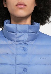 Hele sinine puffer-jakk kõrge krae, õmblusdetailide ja nööpkinnitustega. "GANT" logo rinna piirkonnas. Sile tekstuur.