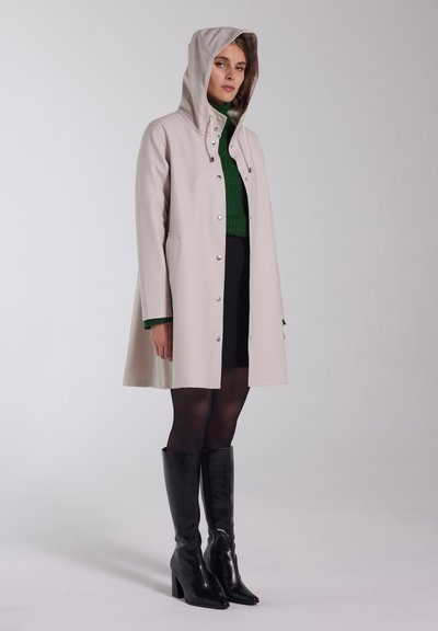 Beige waterdichte jas met een capuchon, drukknoopsluitingen en zijzakken. Gecombineerd met een groene trui, zwarte rok en zwarte kniehoge laarzen.