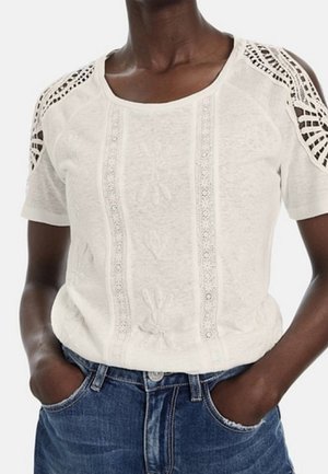 Lysebeige kortærmet top med blomsterblondepaneler foran og hæklet blonde på skuldrene, båret med blå jeans.