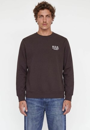 Homme aux cheveux bruns ondulés portant un sweat-shirt marron foncé avec le texte "R.D.D. Royal Denim Division" et un jean bleu sur un fond blanc uni.