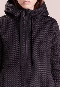 Černá zipová bunda se strukturou v rybí kost, vybavená kapucí a stahovacími šňůrkami, vyrobená z hladkého, lehkého materiálu.