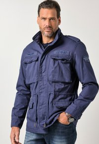 Laivastonsininen takki, jossa on korkea kaulus, useita etutaskuja ja kiristysnyöri vyötäröllä. Takissa on teksturoitu kangas ja kirjailtuja yksityiskohtia.