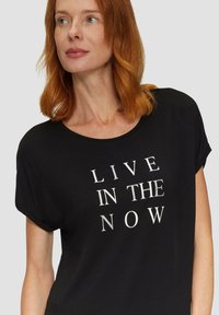 Zwarte T-shirt met korte mouwen en een losse pasvorm, met de tekst "LEEF IN HET MOMENT" in contrasterende witte letters. Zachte stoftextuur.