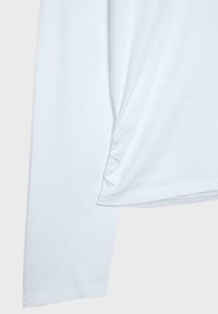 Biała koszula z długim rękawem z marszczonym, marszczonym detalem po boku w pobliżu brzegu, pokazująca fakturę tkaniny i szwy.