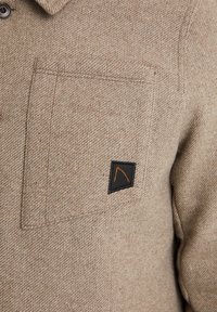 Brun texturerad tygskjorta med en bröstficka som har en svart etikett och orange detaljer. Knappstängning och kragsdesign.