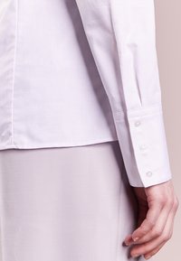 Camicia bianca con un motivo testurizzato; presenta un primo piano del polsino con tre bottoni e una finitura in tessuto liscia e sottile.