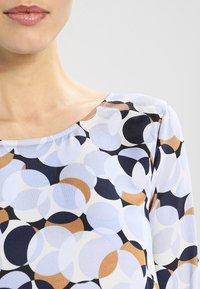 K patterned vršek s kulatými tvary v světle modré, námořnické, hnědé a krémové barvě; texturovaná látka; lodičkový výstřih; tříčtvrteční rukávy.