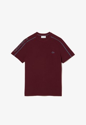 T-shirt bordeaux à manches courtes avec passepoil bleu clair sur les épaules, lettrage Lacoste sur la manche gauche et petit crocodile brodé sur la poitrine.