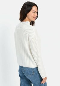 Weißer gerippter Pullover mit hohem Kragen und überschnittenen Schultern, kombiniert mit blauen Jeans. Weiche Textur und lockere Passform, seitlich betrachtet.