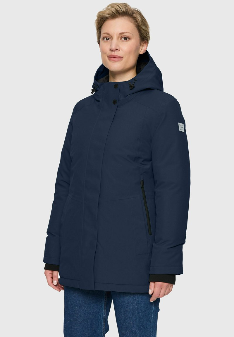 Vrouw in een marineblauwe winterjas met capuchon, voorzien van rits-zijzakken en zwarte manchetten, gecombineerd met een blauwe spijkerbroek, staande tegen een effen achtergrond.