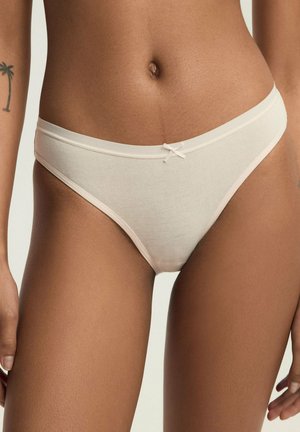 Kobieta nosząca beżowy bawełniany strój kąpielowy z małą kokardką z przodu oraz tatuażem palmowego drzewa na lewym ramieniu.