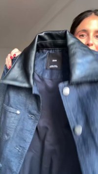 Giubbotto di jeans con colletto in pelle nera, bottoni in metallo argentato e una morbida fodera interna. La combinazione di colori è blu scuro con accenti neri.