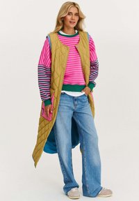 Prešívaná žltá vesta cez ružový prúžkovaný sveter so zelenými akcentmi, v kombinácii s širokými modrými džínsami a sivými teniskami. Výrazný ružový detail na vrecku.