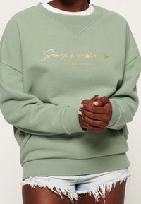 Zielona bluza z okrągłym dekoltem, z złotym logo "Superdry". Noszona z postrzępionymi krótkimi jeansami, prezentującym luźny krój i miękką fakturę.