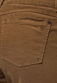 Gros plan d'une poche arrière d'un pantalon en velours côtelé marron avec des coutures visibles et un petit rivet métallique dans le coin supérieur.