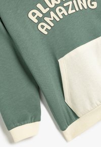 Sudadera verde con un bolsillo crema y puños acanalados. Presenta el texto "SIEMPRE ASOMBROSO" en beige claro. Textura de tela suave.
