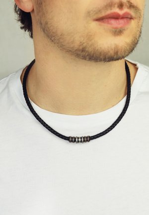 Black braided leather necklace featuring three metallic accent beads in silver and gold tones, worn against a plain white t-shirt.