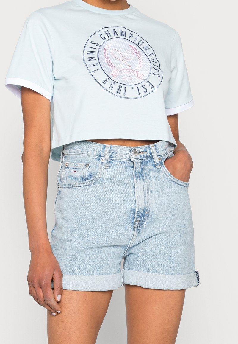 Femme portant un t-shirt court bleu clair avec le logo "Tennis Championships Est. 1999" et un short en jean clair retroussé, main dans la poche.