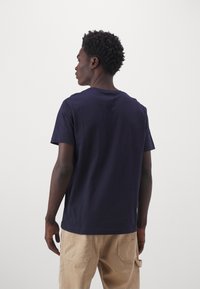 Camiseta de algodón azul marino con cuello redondo y mangas cortas, mostrada por la parte de atrás. Combinada con pantalones beige que muestran detalles de los bolsillos.