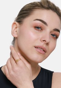 Goldring mit verschlungenem Design, getragen an einer Hand. Model mit heller Haut, blauen Augen und neutralem Make-up, berührt ihr Gesicht. Schwarzes Oberteil.