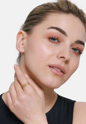 Anello d'oro con design intrecciato, indossato su una mano. Modella con pelle chiara, occhi azzurri e trucco neutro, mentre tocca il suo viso. Maglietta nera.