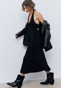 Donna che cammina indossando un vestito halter nero, una giacca di pelle nera poggiata sulle spalle e stivali da cowboy neri contro un muro bianco.