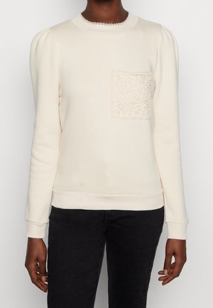 Kvinde iført en cremefarvet langærmet sweater med blonder på kraven og en lomme med blomsterblonde, kombineret med sorte bukser.