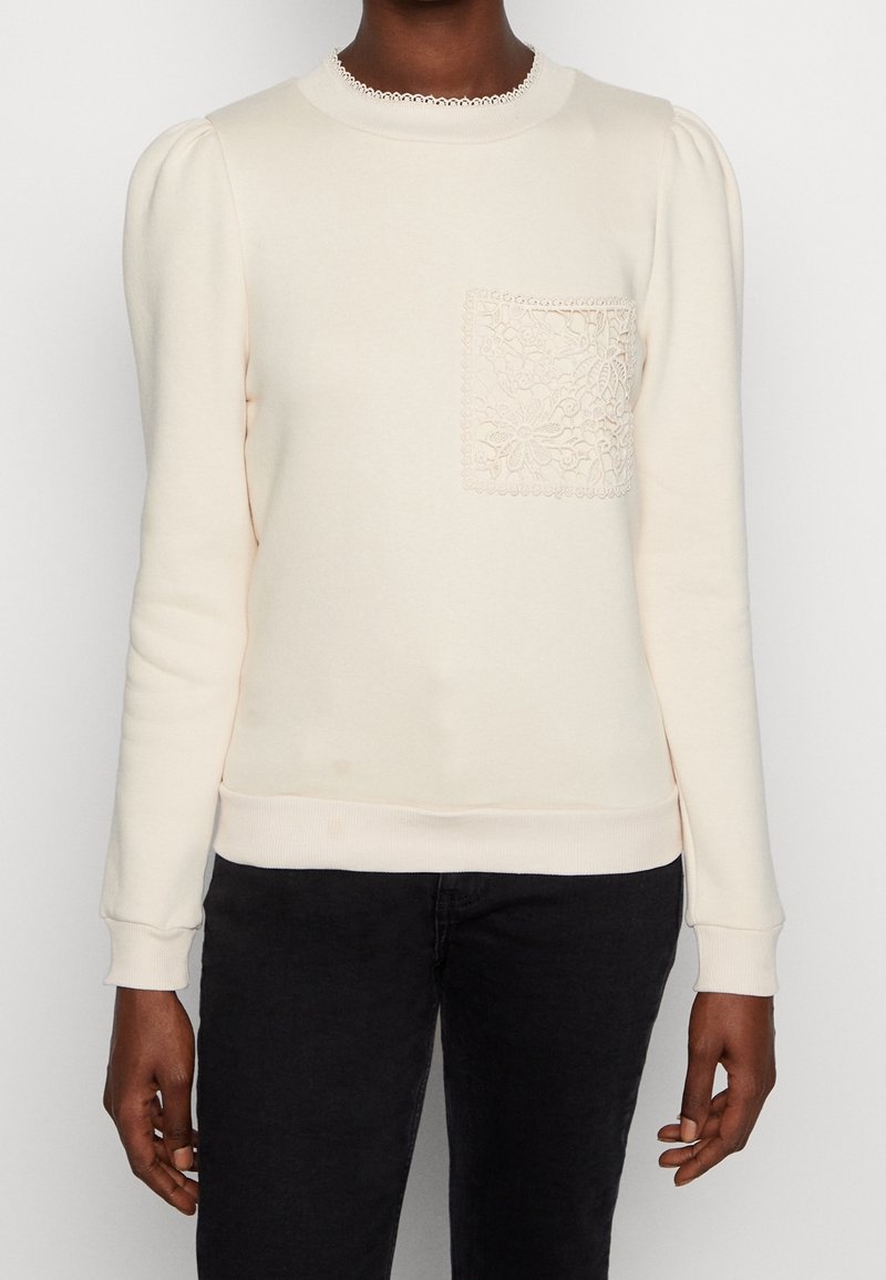 Femme portant un pull crème à manches longues avec un col bordé de dentelle et une poche en dentelle florale, associé à un pantalon noir.