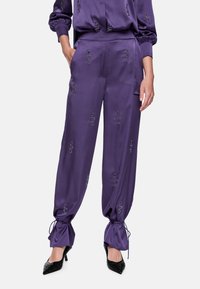 Purple satin pants with embroidered patterns, gathered ankles with ties, side pockets, and worn with black pointed-toe heels.