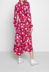 Abito midi floreale con maniche lunghe, caratterizzato da uno sfondo rosa, grandi fiori bianchi e scuri, e una vita annodata. Abbinato a sneakers bianche.