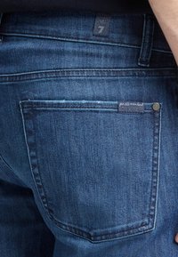 Jeans in denim blu scuro con una tasca posteriore visibile, dotati di un'etichetta tessuta attaccata. La texture del tessuto appare liscia con una leggera scoloritura.