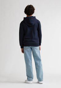 Marineblauwe capuchontrui, losse pasvorm, gecombineerd met lichtblauwe losse jeans. Witte sneakers zichtbaar, achterkant zicht. Gladde stoftextuur.