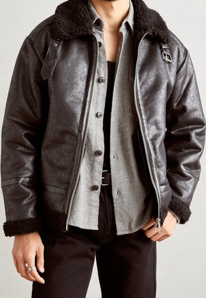 Mann trägt eine schwarze Lammfell-Lederjacke über einem grauen Hemd mit Knöpfen und schwarzen Hosen, mit einer Hand in der Tasche und einem silbernen Ring am Finger.