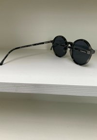 Lunettes de soleil rondes marbrées noires et grises avec des verres sombres, monture en acétate transparent et branches courbées. Logo de la marque sur le côté.