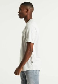 T-shirt di cotone grigio chiaro con maniche corte, fessure laterali e un piccolo logo sul petto, abbinato a jeans blu chiaro.