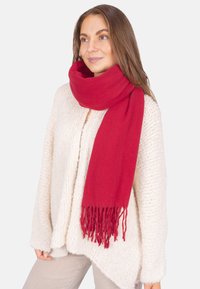 Roter Schal aus weichstoff, mit fransigen Kanten, über einem cremefarbenen, strukturierten Pullover getragen. Einfaches und warmes Design.