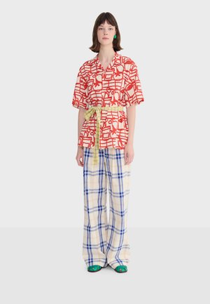 Frau trägt ein kurzärmliges Hemd mit rotem und weißem Muster, einen gelben Gürtel, weit geschnittene blau-beige karierte Hosen und grüne Sandalen.