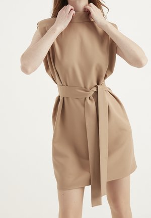 Woman wearing sleeveless beige dress with wide belt tied at the waist, adjusting collar, against plain white background.