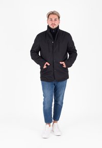 Giacca nera con colletto alto e bottoni a pressione, dotata di due tasche con zip e una texture liscia, abbinata a jeans blu e sneaker bianche.
