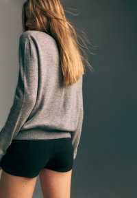 Donna con lunghi capelli biondi, che indossa un maglione grigio e pantaloni corti neri, girata di profilo su uno sfondo grigio scuro.